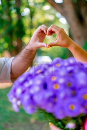 Photo for Close-up of a young couple's hands joining and showing a heart next to a bouquet of flowers in park - Royalty Free Image