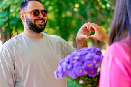 Foto de Primer plano de las manos de una joven pareja uniéndose y mostrando un corazón junto a un ramo de flores en el parque - Imagen libre de derechos