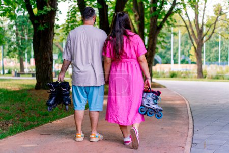 Foto de Una joven pareja camina por el parque mientras está en una cita cogida de la mano y sosteniendo patines en las manos de modelos de talla grande - Imagen libre de derechos