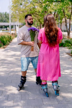 Photo for Young couple on roller skates guy gives girl a bouquet of flowers during date in the park - Royalty Free Image