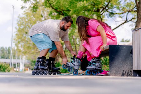 Photo for Young couple guy helps girl put on rollerblades who is sitting on a bench in the park while dating plus size models - Royalty Free Image