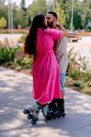 Photo for A young couple on rollerblades kiss during a date in the park against backdrop of trees - Royalty Free Image