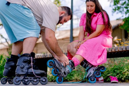 Foto de Joven pareja chico ayuda a chica poner en patines que está sentado en un banco en el parque mientras citas más modelos de tamaño - Imagen libre de derechos