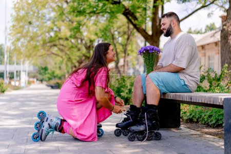 Téléchargez les photos : Jeune couple fille aide gars mis sur rollerblades qui est assis sur un banc de parc tenant un bouquet tout en datant modèles plus la taille - en image libre de droit