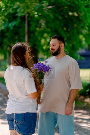 Téléchargez les photos : Jeune couple gai gars donnant des fleurs à la fille dans le parc pendant la date sur fond d'arbres plus les modèles de taille - en image libre de droit