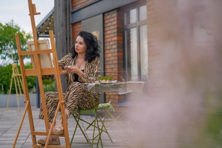 Téléchargez les photos : Jeune jolie fille artiste assis sur une chaise dans la rue et avec un pinceau peint une image sur toile debout sur un chevalet - en image libre de droit