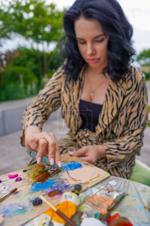 Téléchargez les photos : Jeune jolie fille artiste assis sur une chaise dans la rue et prend de la peinture de la palette avec peinture spatule - en image libre de droit