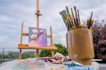 Téléchargez les photos : Gros plan d'un stand de peinture avec pinceaux contre la rue et peinture sur un chevalet - en image libre de droit