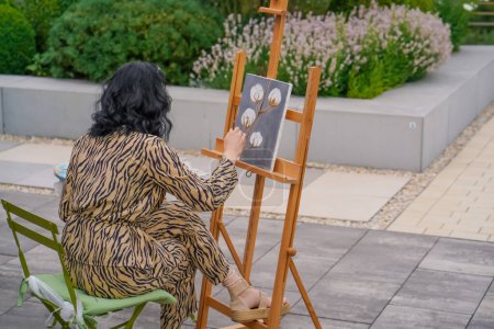 Téléchargez les photos : Une jeune fille artiste peint le tableau avec un pinceau sur une toile qui se dresse sur un chevalet vue de derrière le dos - en image libre de droit