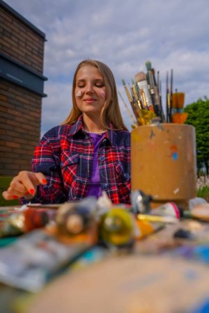 Téléchargez les photos : Portrait de joyeuse fille artiste doigt peinture sur son visage assis à une table de peinture avec des tubes de peinture et des pinceaux sur elle - en image libre de droit