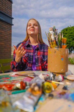 Téléchargez les photos : Portrait de joyeuse fille artiste doigt peinture sur son visage assis à une table de peinture avec des tubes de peinture et des pinceaux sur elle - en image libre de droit