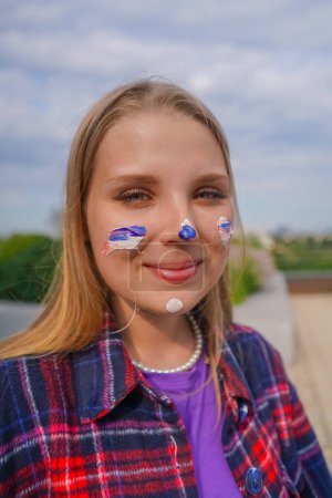 Foto de Retrato de una joven artista alegre con pintura al óleo en la cara sonriendo en el fondo de la ciudad - Imagen libre de derechos
