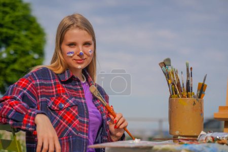 Téléchargez les photos : Portrait d'une jeune fille artiste joyeuse avec de la peinture à l'huile sur son visage et des pinceaux dans ses mains souriant sur fond de ville - en image libre de droit
