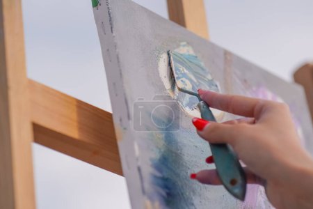 Photo for A close-up of a painting spatula applying oil paint to a painting that is standing on an easel in the process of creating painting - Royalty Free Image