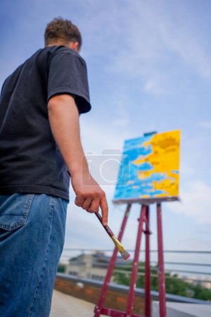 Foto de Un joven artista se para con un pincel en la mano delante de tres pinturas paradas en caballetes vista desde detrás de él - Imagen libre de derechos