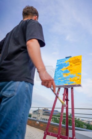 Téléchargez les photos : Un jeune artiste se tient avec un pinceau à la main devant trois tableaux debout sur des chevalets vue de derrière lui - en image libre de droit