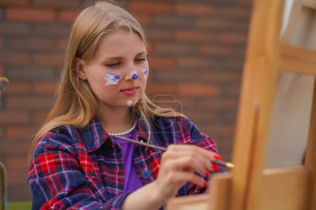 Téléchargez les photos : Focalisée fille artiste avec de la peinture sur son visage peinture une image debout sur un chevalet avec pinceau - en image libre de droit