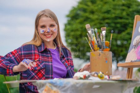 Foto de Retrato de una artista alegre con pintura al óleo en la cara y pinceles en las manos sonriendo sobre el fondo de la ciudad - Imagen libre de derechos