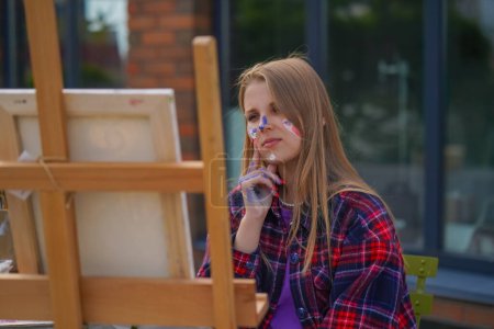 Téléchargez les photos : Focalisée fille artiste avec de la peinture sur son visage peinture une image debout sur un chevalet avec pinceau - en image libre de droit