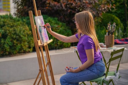 Téléchargez les photos : Une jeune fille artiste peint un tableau avec un pinceau sur une toile qui se tient sur chevalet vue de derrière le dos - en image libre de droit