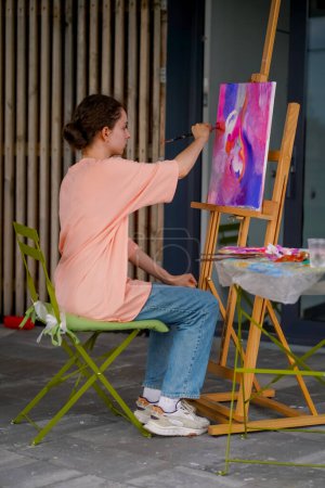 Photo for A girl artist with problem skin sits at an easel and paints a picture with brush in her studio - Royalty Free Image