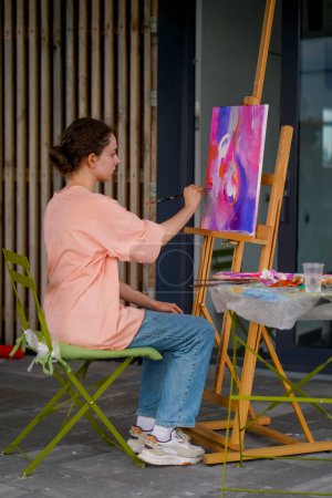 Téléchargez les photos : Une fille artiste avec une peau problématique s'assoit à un chevalet et peint un tableau avec un pinceau dans son atelier - en image libre de droit