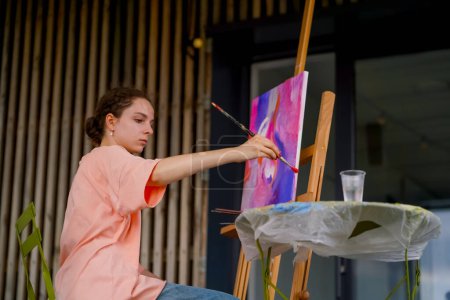 Foto de Una joven artista con piel problemática se sienta cerca de un caballete y toma pintura de la paleta de dibujo - Imagen libre de derechos
