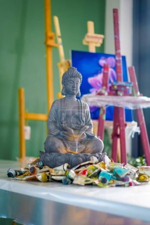 Téléchargez les photos : Un gros plan de la statue de la fontaine de Bouddha debout sur une table dans un atelier de peinture avec des tubes de peinture à côté - en image libre de droit