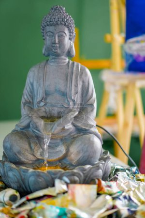 Foto de Un primer plano de la estatua de la fuente de Buda de pie sobre una mesa en un estudio de pintura con tubos de pintura al lado - Imagen libre de derechos