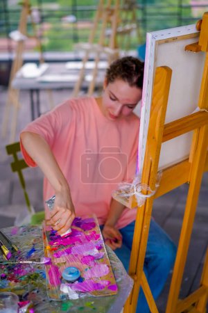 Téléchargez les photos : Gros plan d'une fille artiste ayant un problème de peau pressant de la peinture d'un tube sur une palette tout en peignant - en image libre de droit