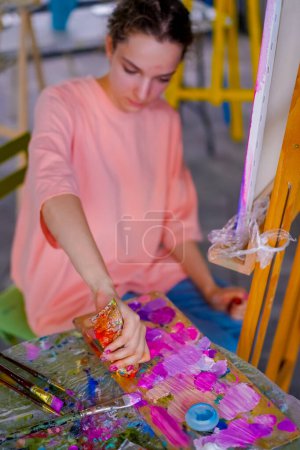 Téléchargez les photos : Gros plan d'une fille artiste ayant un problème de peau pressant de la peinture d'un tube sur une palette tout en peignant - en image libre de droit