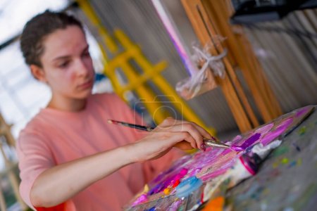 Foto de Una joven artista con piel problemática se sienta cerca de un caballete y toma pintura de la paleta de dibujo - Imagen libre de derechos