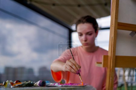 Téléchargez les photos : Une jeune artiste féminine avec une peau problématique s'assoit près d'un chevalet et prend de la peinture de la palette de dessin - en image libre de droit