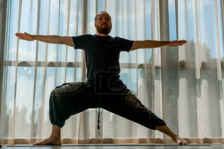 Photo for A male yogi stands in a warrior pose from kundalini yoga in sunny gym for training hobby active lifestyle - Royalty Free Image