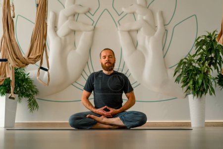 Téléchargez les photos : Un homme adulte avec une barbe tient ses mains devant lui et médite les yeux fermés en position lotus dans un studio de yoga léger - en image libre de droit