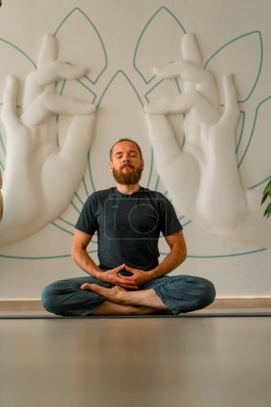 Foto de Un hombre adulto con barba se sienta y medita con los ojos cerrados en posición de loto en un luminoso estudio de yoga - Imagen libre de derechos