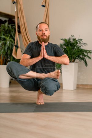 Téléchargez les photos : Concentré yogi mâle adulte avec une barbe effectue un exercice de pose de yoga médite et maintient l'équilibre dans le hall le fond des fleurs - en image libre de droit
