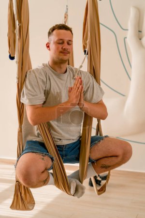 Téléchargez les photos : Un gars souriant joyeux accroche sur des toiles pour le yoga de mouche dans la salle de yoga et regarde dans le cadre - en image libre de droit