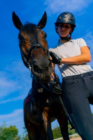 Photo for A horsewoman in a helmet hugs and scratches her beautiful black horse with her hand at the equestrian arena during horseback ride - Royalty Free Image
