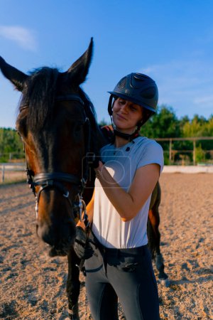 Foto de Una jinete en un casco abraza y rasca su hermoso caballo negro con su mano en la arena ecuestre durante el paseo a caballo - Imagen libre de derechos