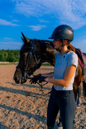 Foto de Un jinete con casco alimenta a su hermoso caballo negro de su mano en la arena ecuestre durante el paseo a caballo - Imagen libre de derechos