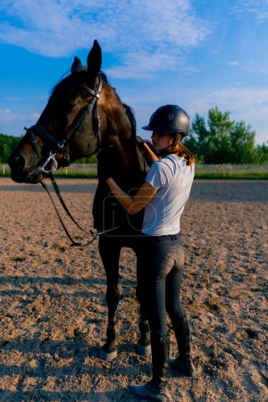 Foto de Un jinete de casco lleva a su hermoso caballo negro por el arnés en la arena de montar a caballo durante el paseo - Imagen libre de derechos