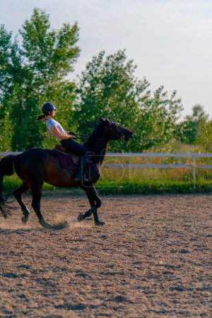Foto de Una jinete vestida con un casco monta su hermoso caballo negro en una arena para montar a caballo durante el paseo a caballo - Imagen libre de derechos