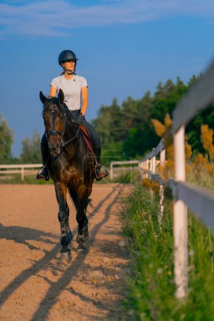 Foto de Una jinete vestida con un casco monta su hermoso caballo negro en una arena para montar a caballo durante el paseo a caballo - Imagen libre de derechos