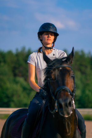 Téléchargez les photos : Une cavalière casquée conduit son beau cheval noir par le harnais dans l'arène d'équitation pendant la randonnée à cheval - en image libre de droit