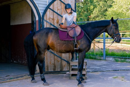 Foto de Un jinete con casco monta su hermoso caballo negro cerca del establo antes de un paseo a caballo en el bosque - Imagen libre de derechos