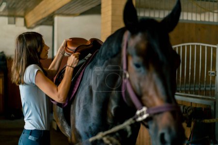 Téléchargez les photos : Une cavalière met sa selle sur son cheval noir dans les écuries en préparation du concept équestre de course - en image libre de droit