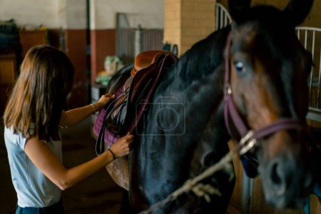 Photo for A rider puts saddle on her black horse in the stables in preparation for the race equestrian concept - Royalty Free Image