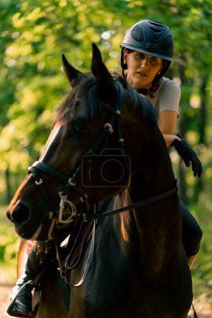 Téléchargez les photos : Une cavalière vêtue d'un casque monte son beau cheval noir dans la forêt lors d'une balade à cheval - en image libre de droit