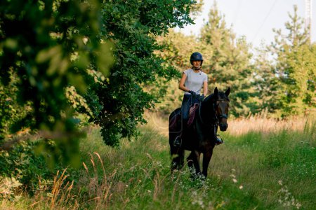 Foto de Una jinete vestida con un casco monta su hermoso caballo negro en el bosque durante el paseo a caballo - Imagen libre de derechos